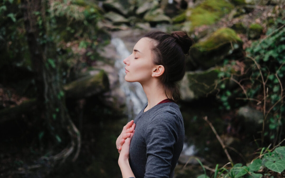 Young woman practicing breathing yoga pranayama outdoors in moss forest on background of waterfall. Unity with nature concept.